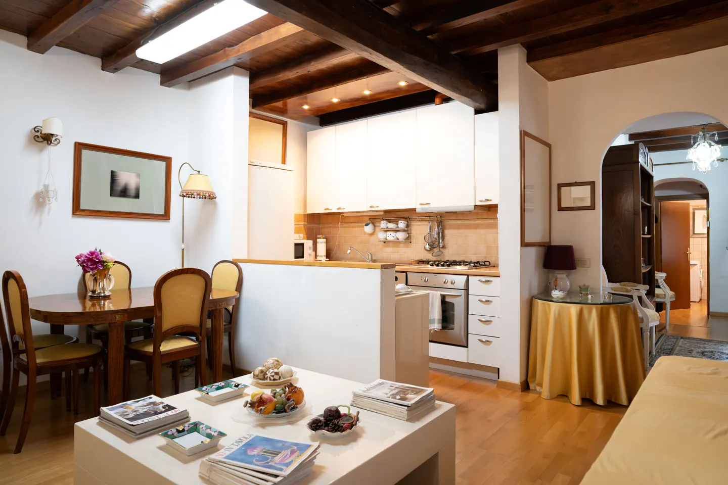 Open-concept living space with wood floors, exposed beams, and white walls. A dining table, kitchen, and living area are visible.