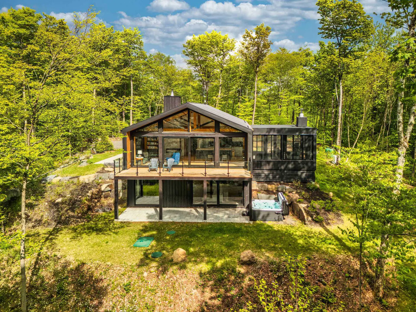 Modern black cabin with a glass-walled deck, hot tub, and patio, nestled in a lush green forest under a blue sky.
