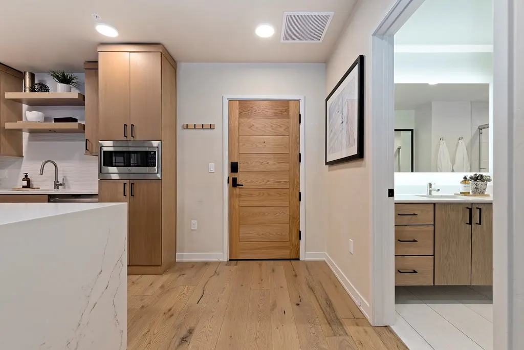Interior view of a modern apartment with light wood floors, cabinets, and a front door. A bathroom is visible through a doorway.