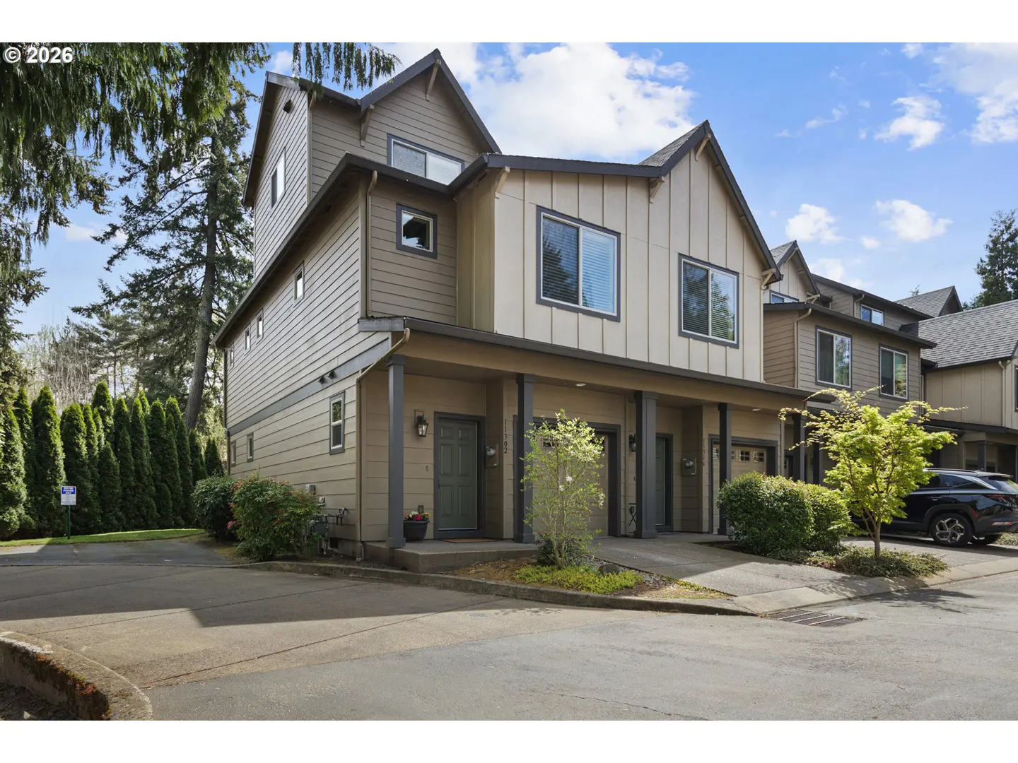 A three-story townhouse with beige siding, dark trim, and a covered porch. A driveway leads to the garage. Green trees surround the building.