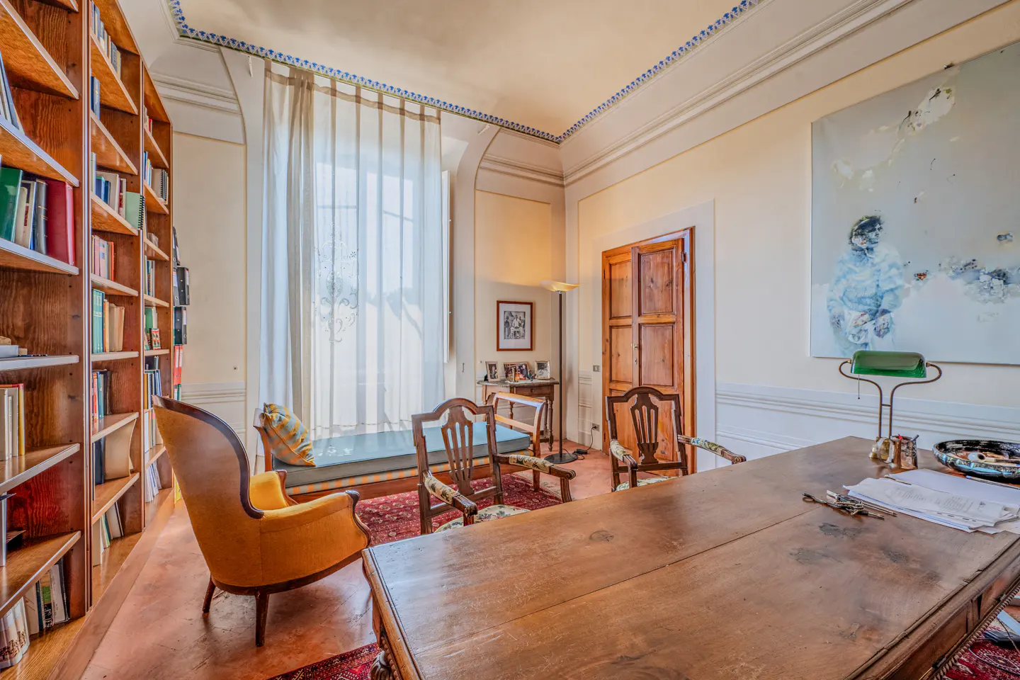 A home office with a large wooden desk, yellow armchair, two wooden chairs, a daybed by a window, and a large wall painting.