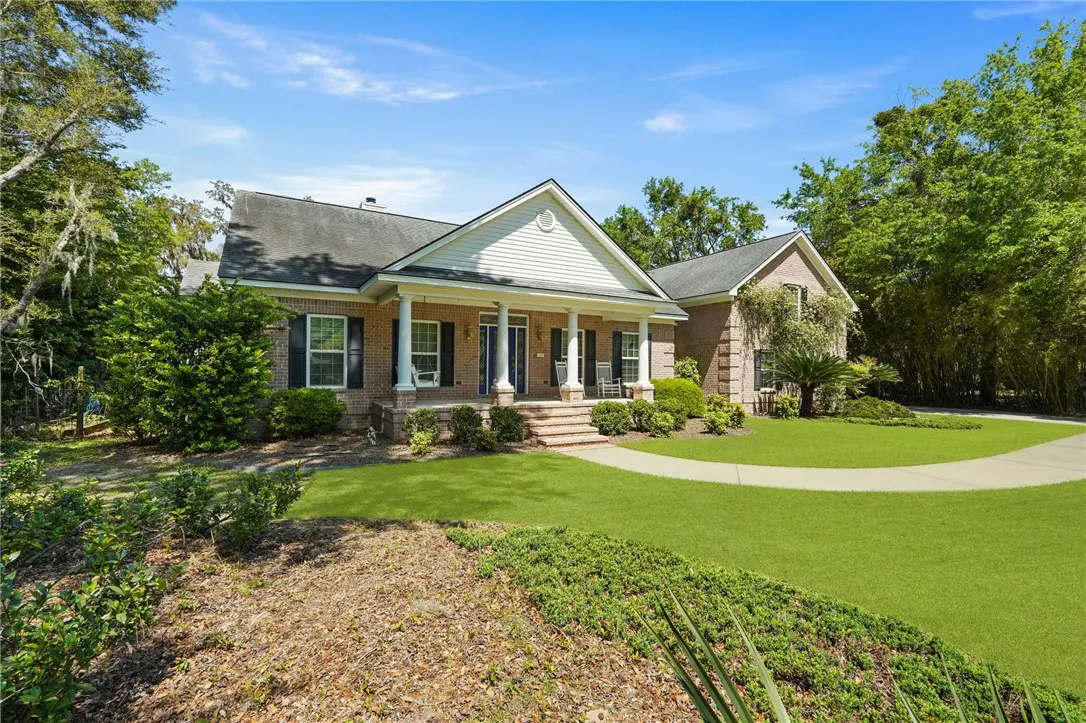 Brick house with white trim, black shutters, and a green lawn under a blue sky. Trees surround the property.