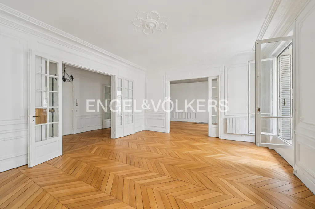 Bright, empty room with herringbone wood floors, white walls, and open glass doors. Ceiling has decorative molding.