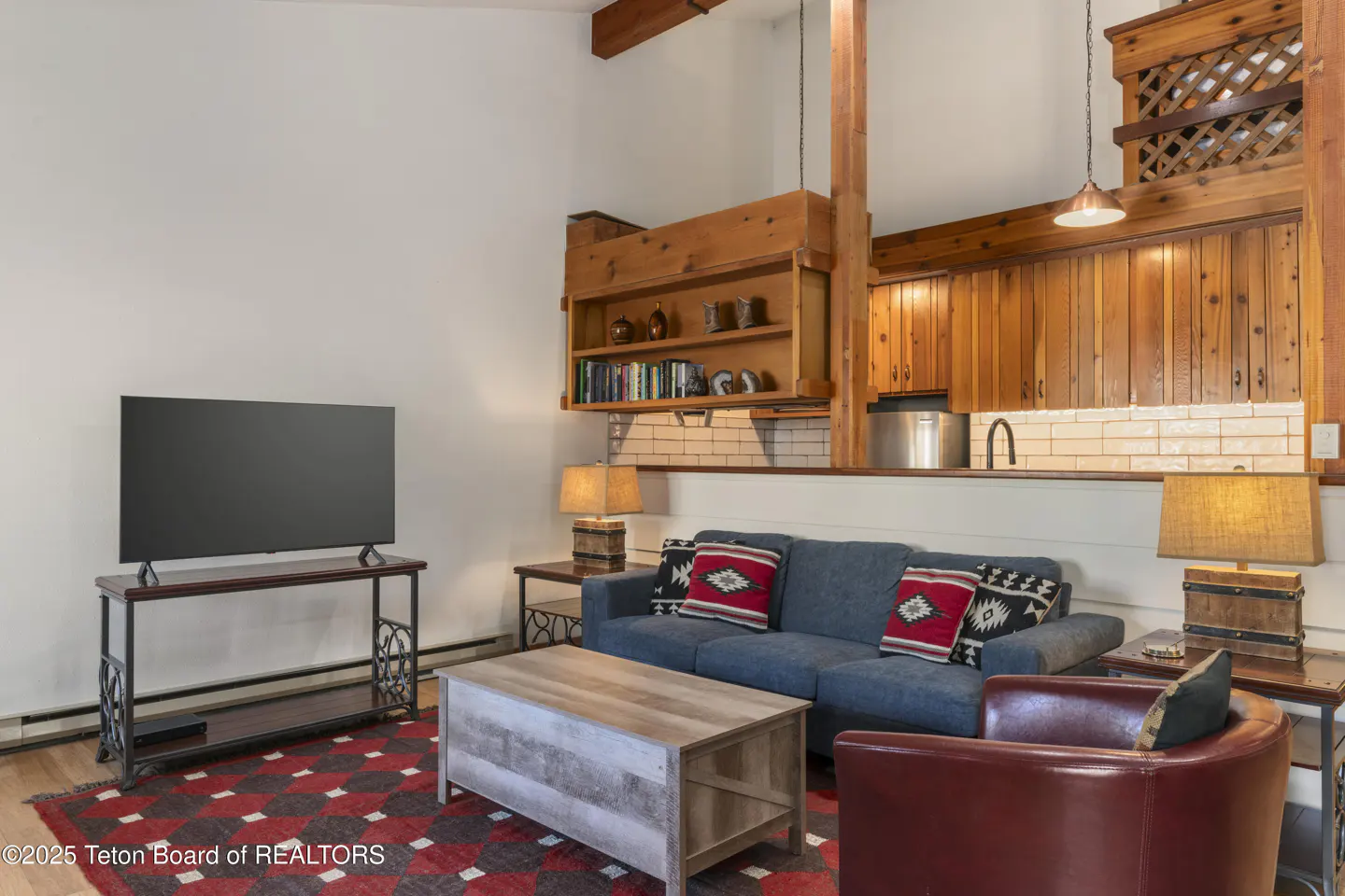 Living room with blue sofa, red patterned rug, TV, and wood cabinets above a white tiled backsplash.