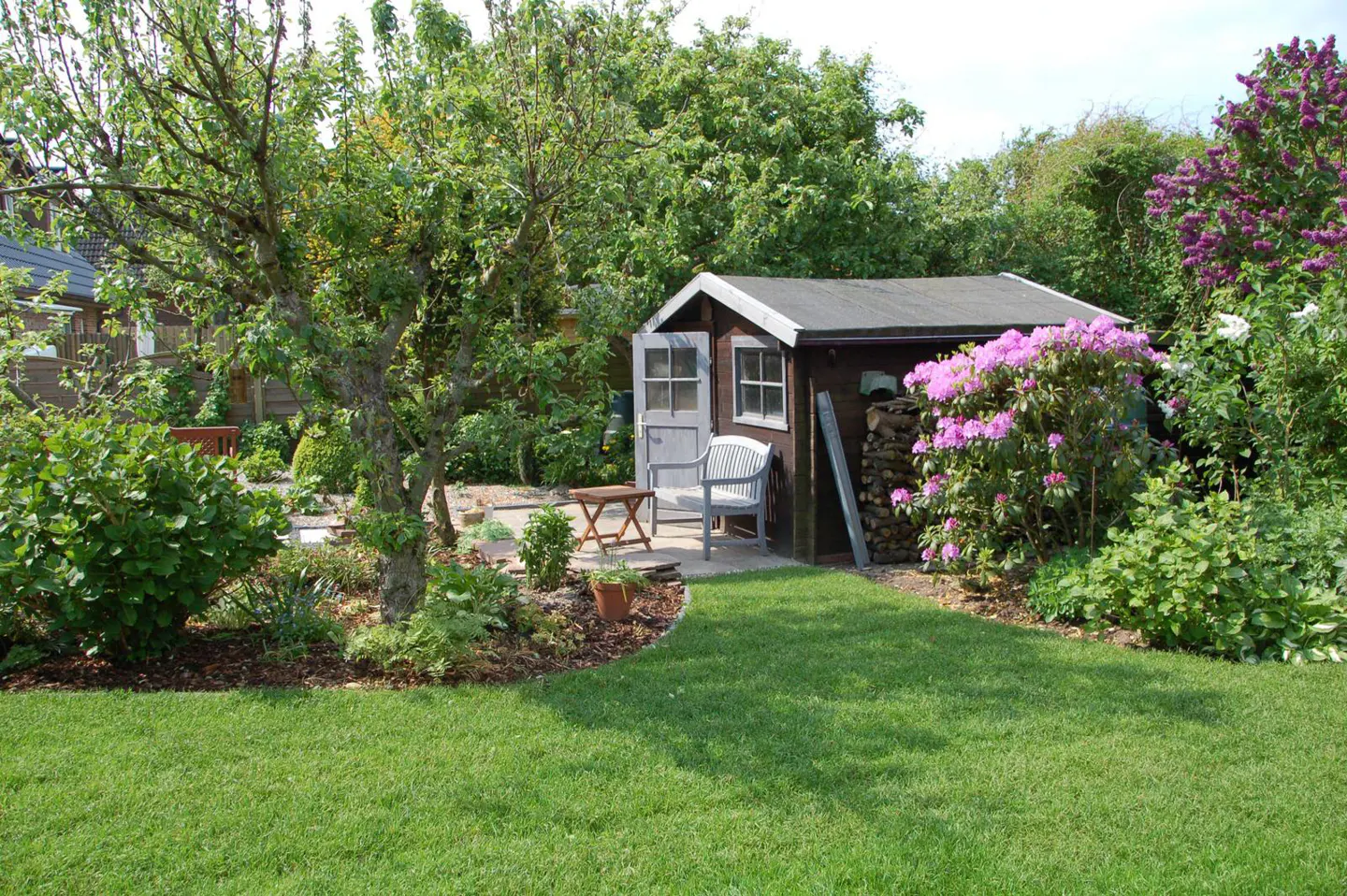 A backyard with a shed, green grass, trees, and flowers. A chair and table sit outside the shed.