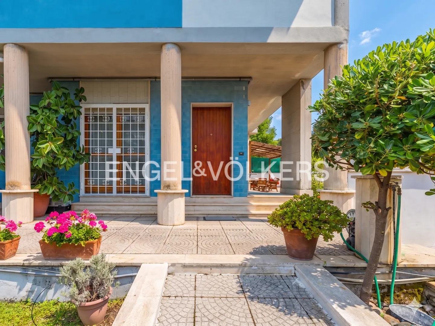 Exterior of a blue and white house with a brown front door, columns, and potted plants. The Engel & Völkers logo is visible.