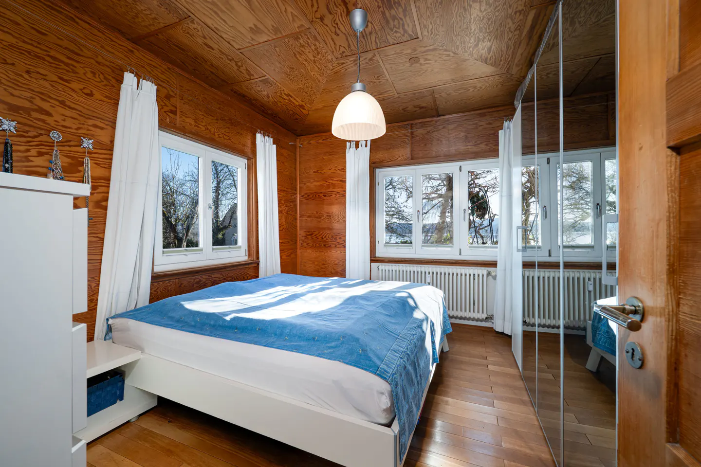 Bedroom with wood-paneled walls, white bed with blue cover, and mirrored closet. Windows show trees.