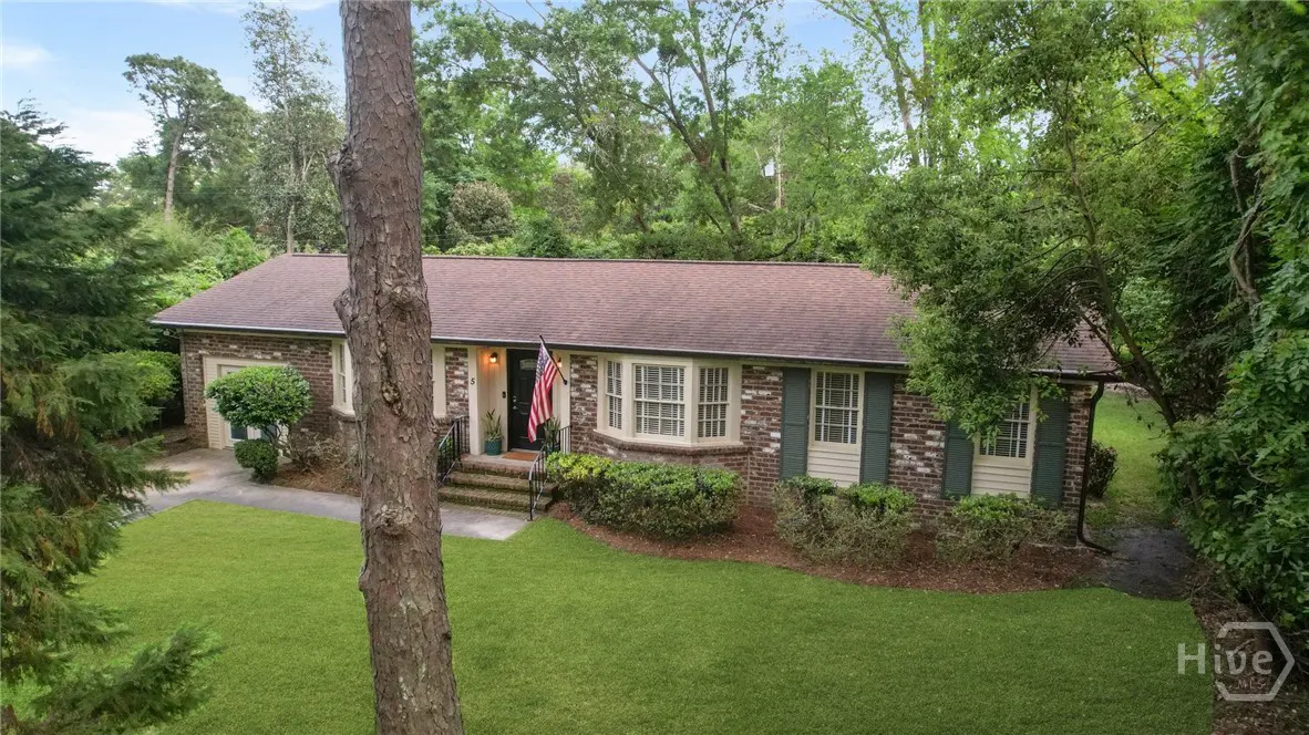 A single-story brick house with green shutters and a brown roof, surrounded by green trees and grass. An American flag hangs near the front door.