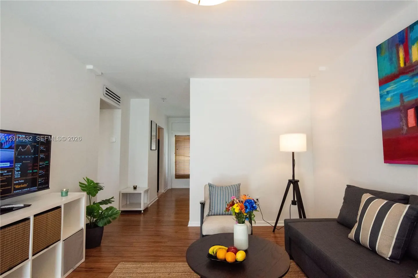 Bright living room with hardwood floors, white walls, and a gray sofa. A colorful painting hangs above the sofa. A TV displays stock market data.