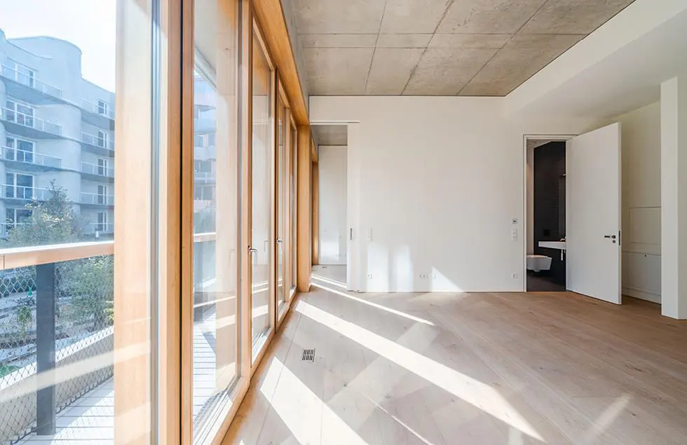 Bright, empty room with wood floors, white walls, and a concrete ceiling. Large windows with wood frames overlook a balcony and building. Open bathroom door.