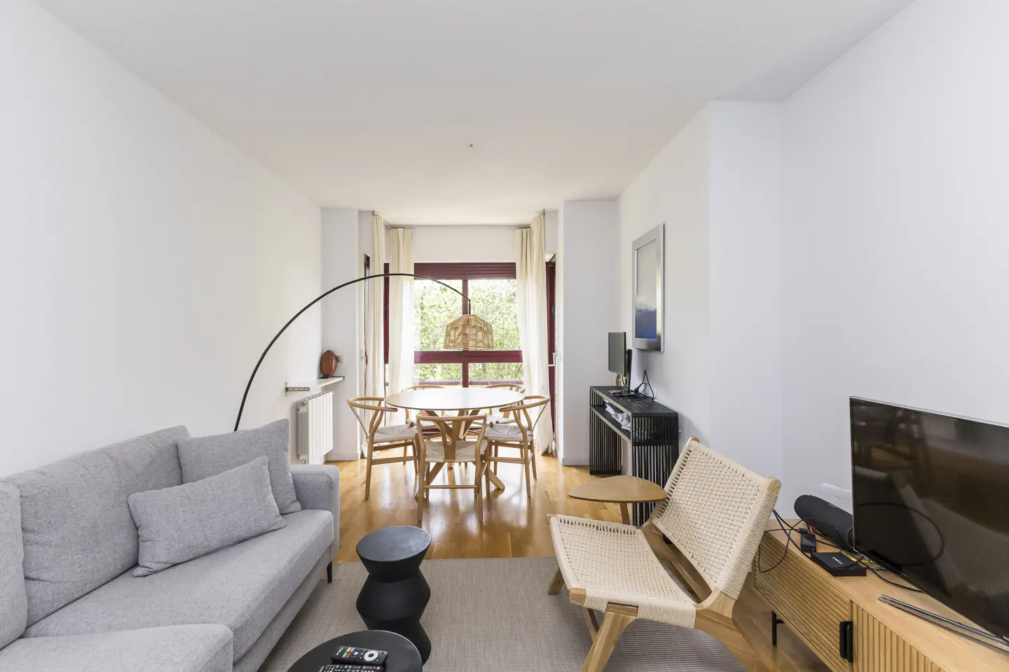 Bright living room with a gray sofa, round wood table with chairs, and a woven chair. Hardwood floors and white walls.