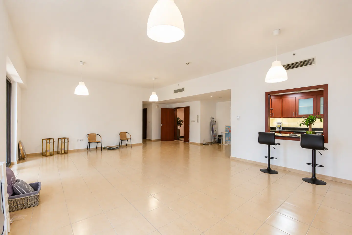 Bright, empty living room with beige tile floor, white walls, and pendant lights. Kitchen bar with two stools visible.
