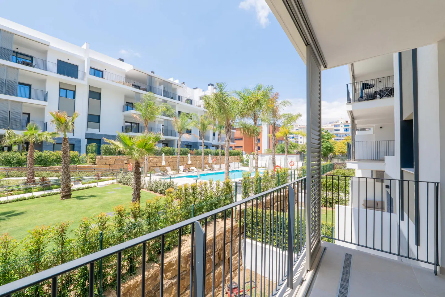 View from a balcony overlooking a modern apartment complex with a swimming pool, palm trees, and a manicured green lawn.