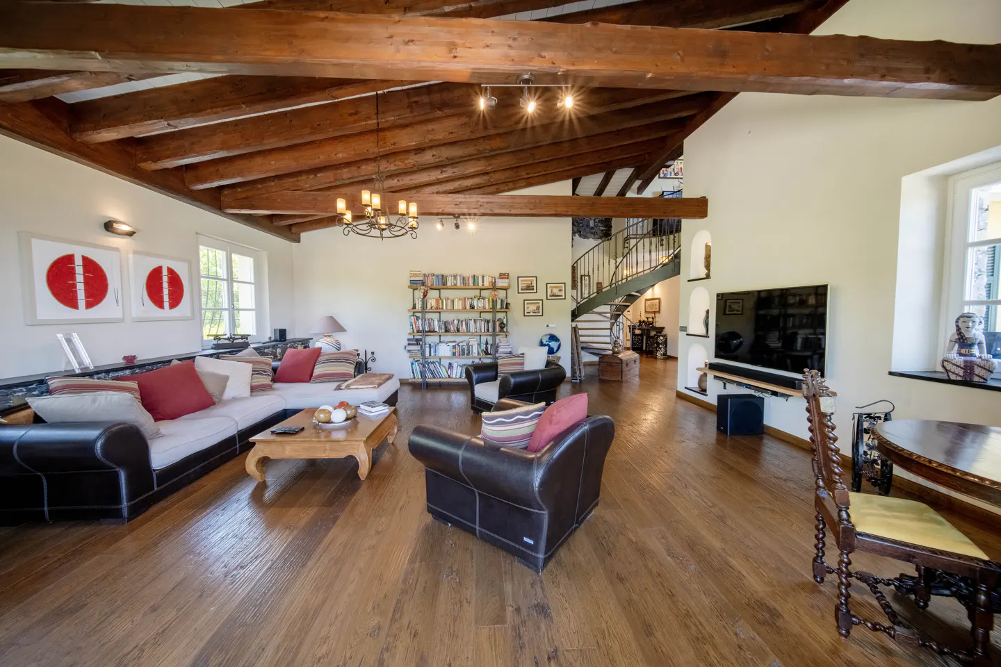 A spacious living room with hardwood floors, exposed wood beams, and a spiral staircase. A large sectional sofa and leather armchairs provide seating.