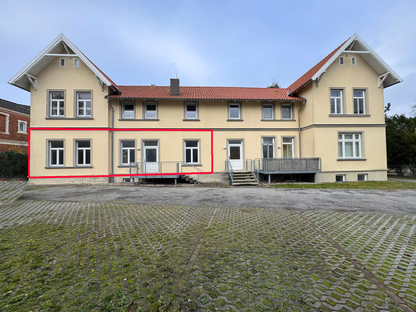 Rear view of a yellow two-story building with a red outline, showing windows, doors, and a gray stone parking lot.