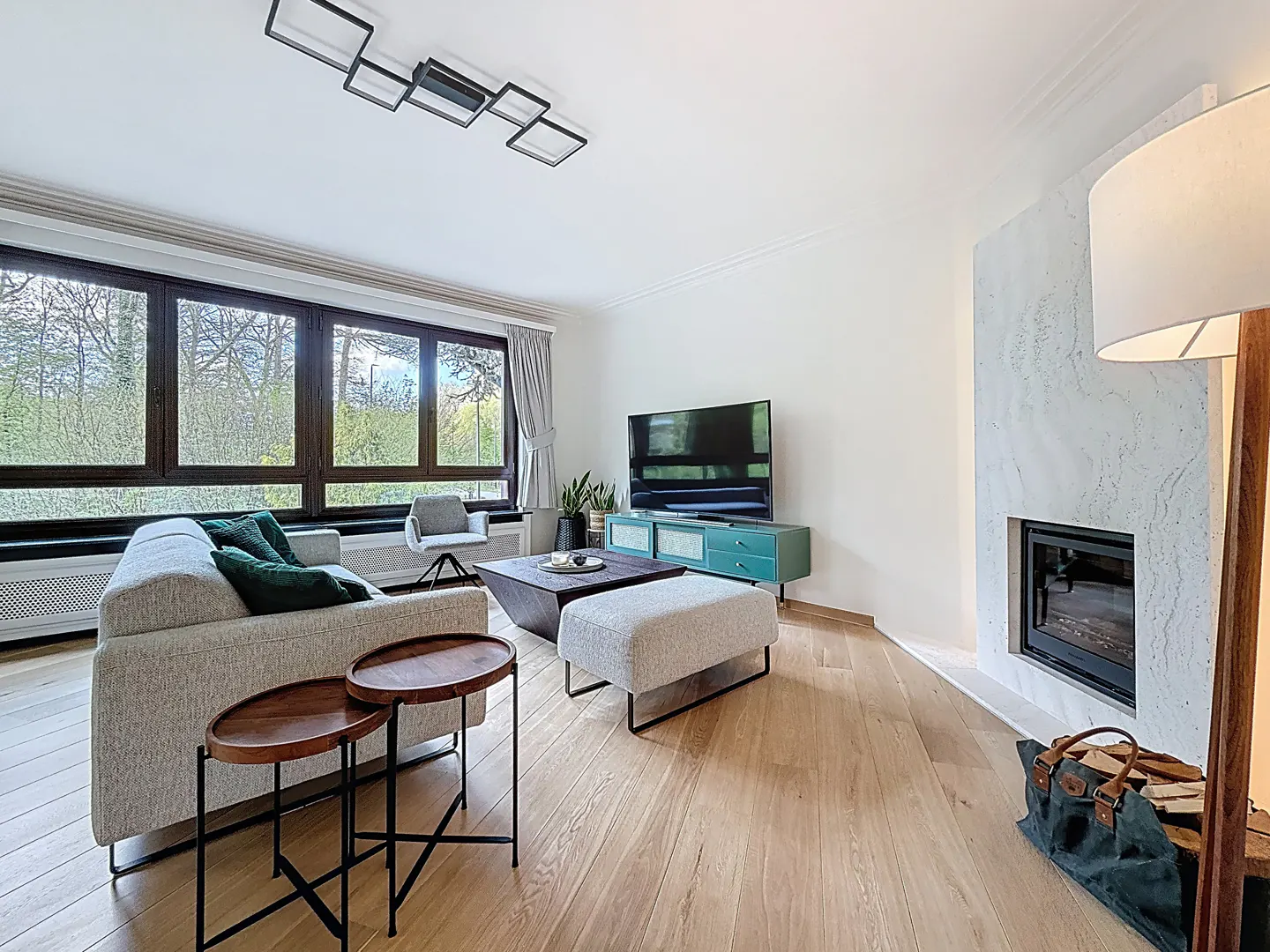 Bright living room with wood floors, a gray sofa with green pillows, and a modern fireplace. Large windows overlook a green landscape.