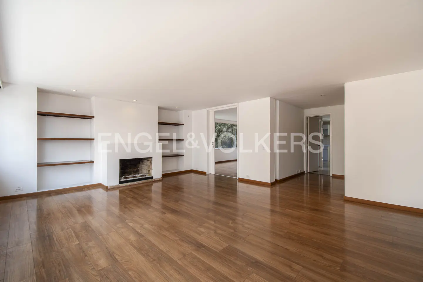 Bright, empty living room with hardwood floors, white walls, fireplace, and built-in shelves. Doorways lead to other rooms.