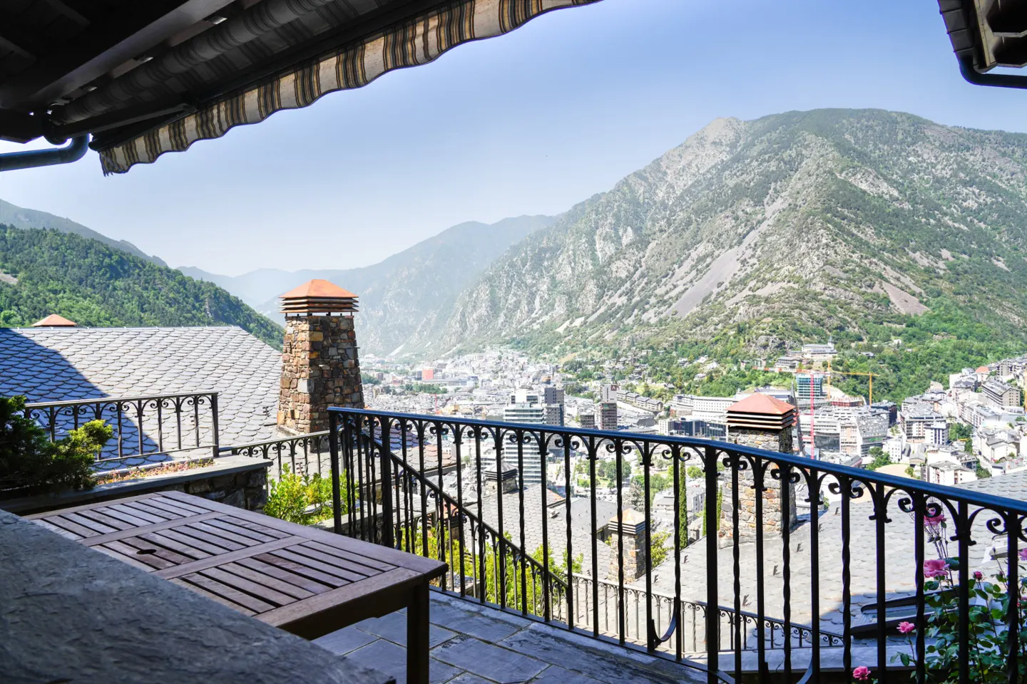 View from a balcony with a wooden table and black iron railing overlooking a city nestled in a valley surrounded by mountains.
