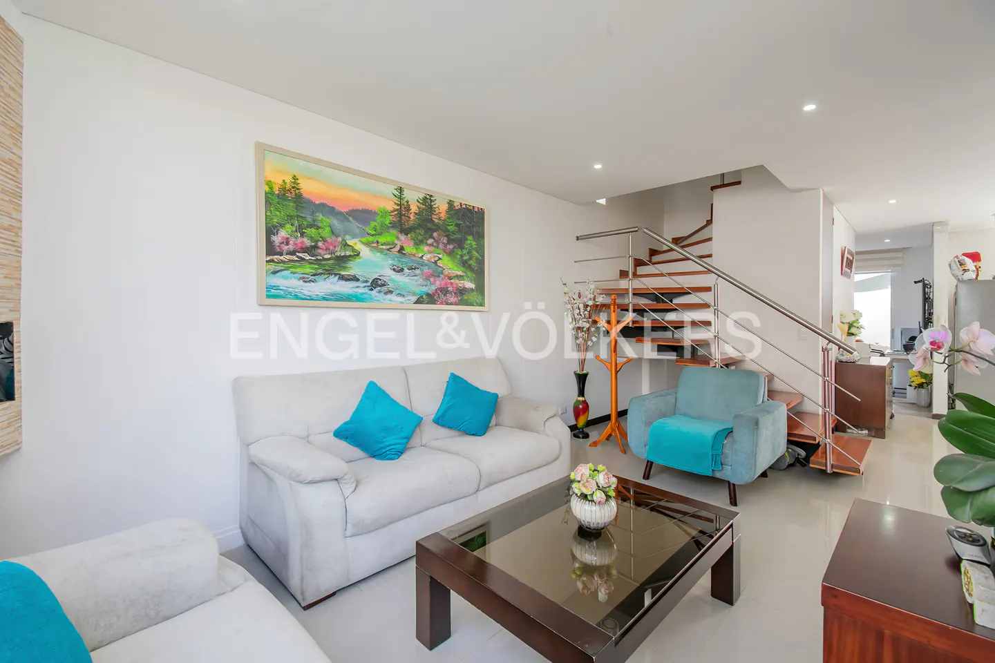 Living room with white walls, light gray sofas with blue pillows, a painting, and a wooden staircase.