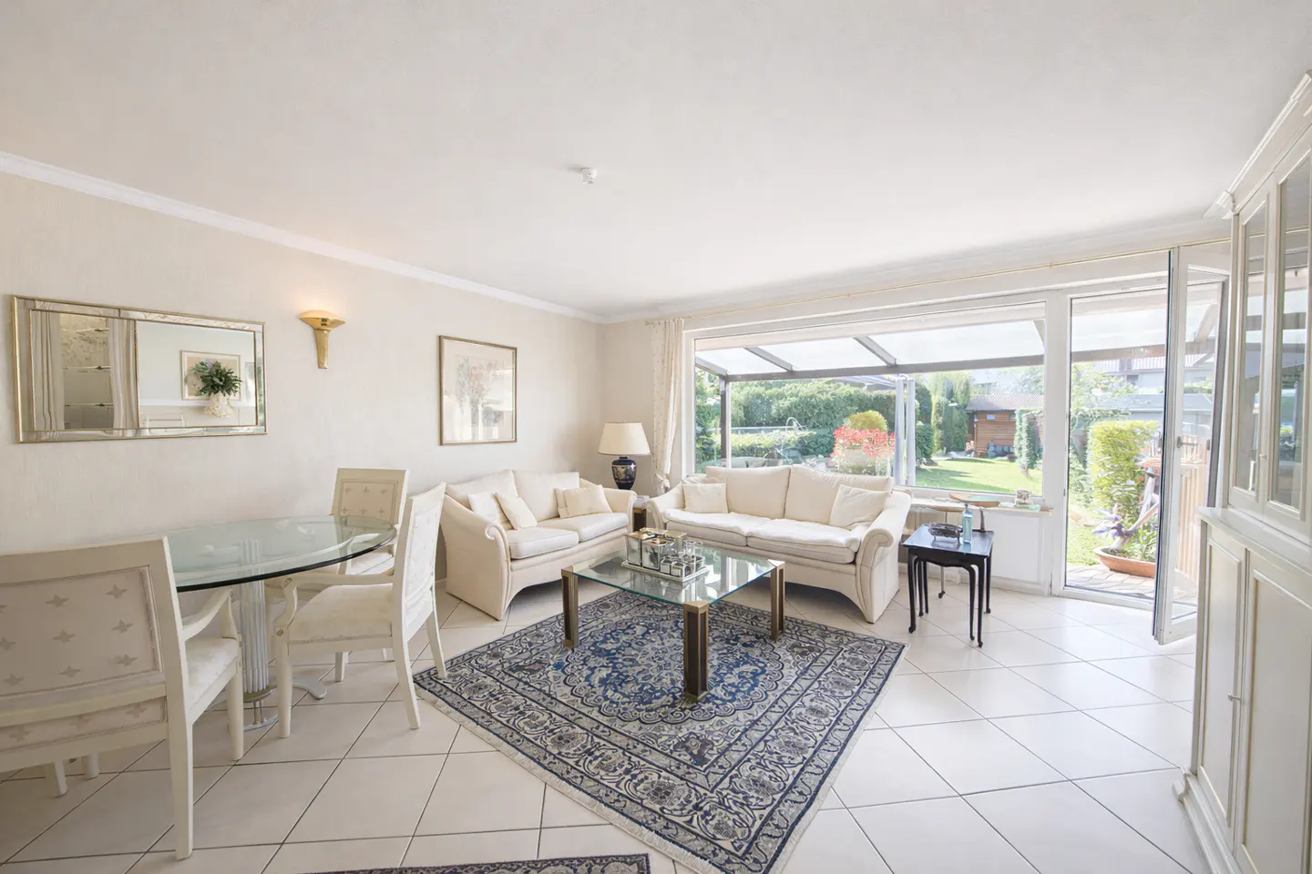 Bright living room with white tile floors, two white sofas, a glass table, and a blue rug. A glass door leads to a green garden.