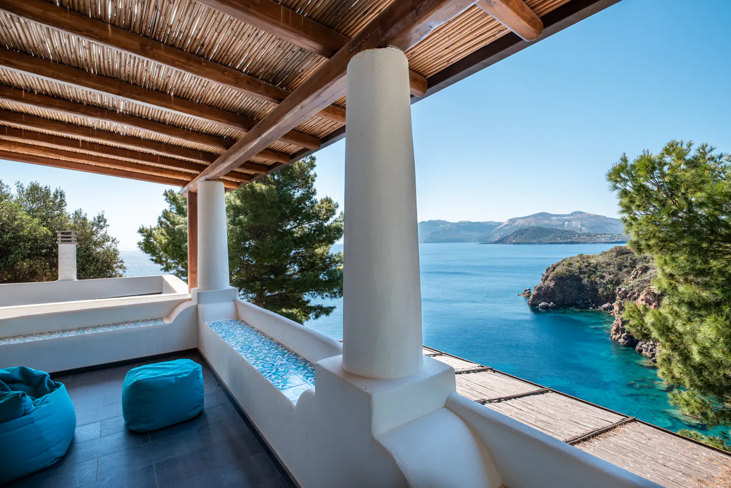 Balcony with blue beanbag chairs, white columns, and a thatched roof overlooking the blue sea and green islands on a sunny day.