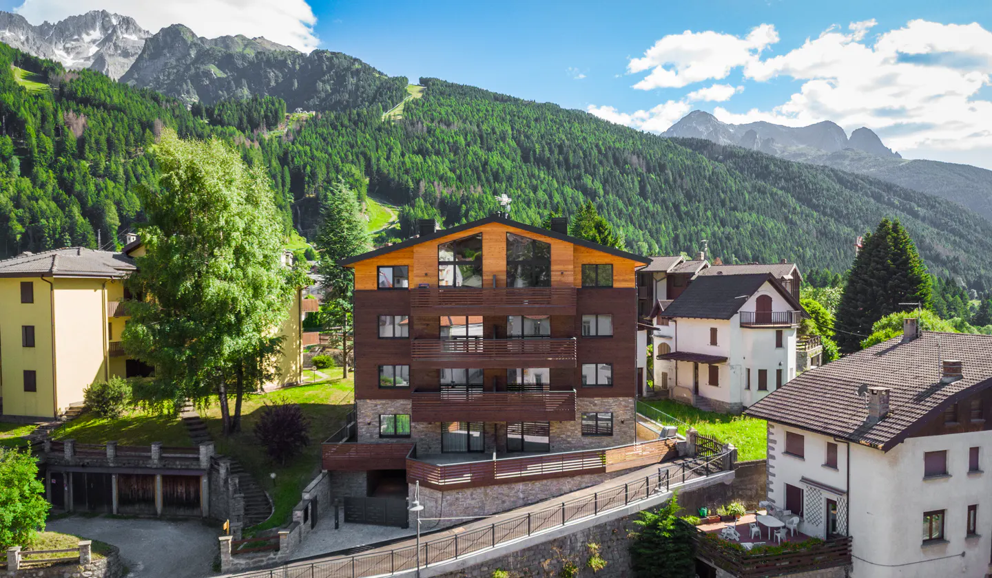 A three-story brown and tan building with balconies, nestled in a green, mountainous landscape under a blue sky.