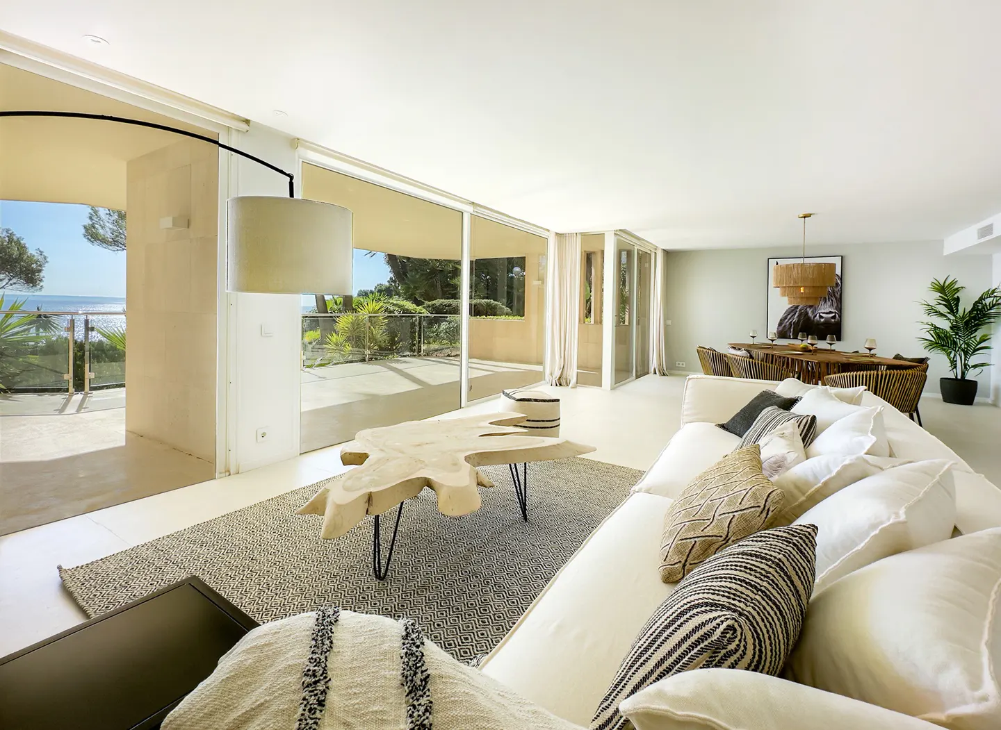 Bright living room with white sofa, patterned pillows, and a unique wood coffee table. Large windows offer ocean views. Dining area in background.