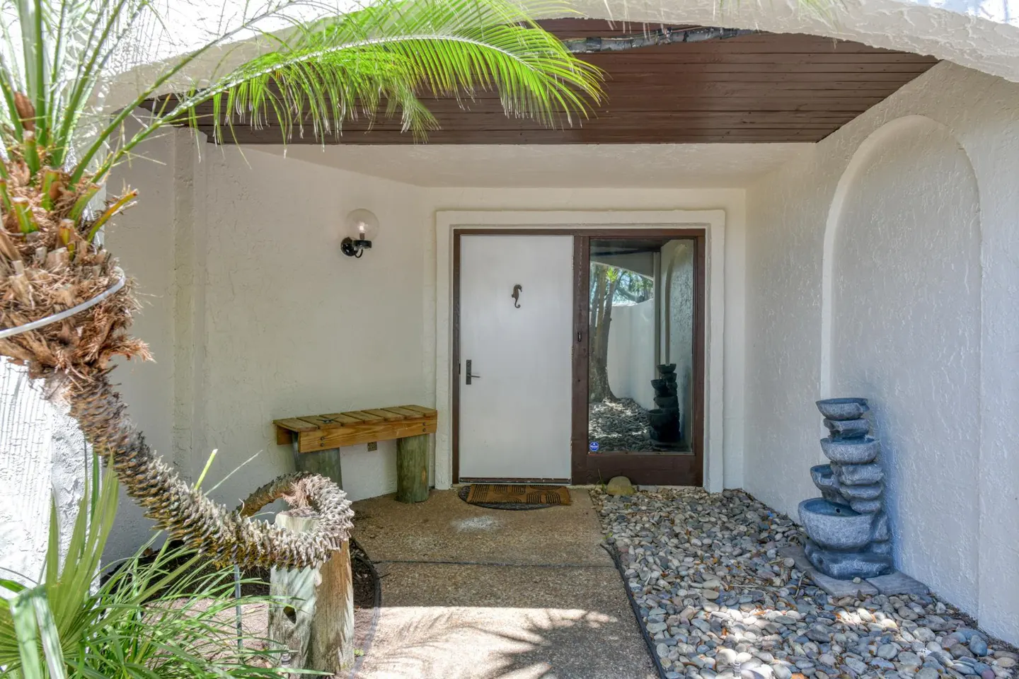 Home's front entrance with a white door, glass-paneled door, and a small water fountain. A palm tree is on the left.