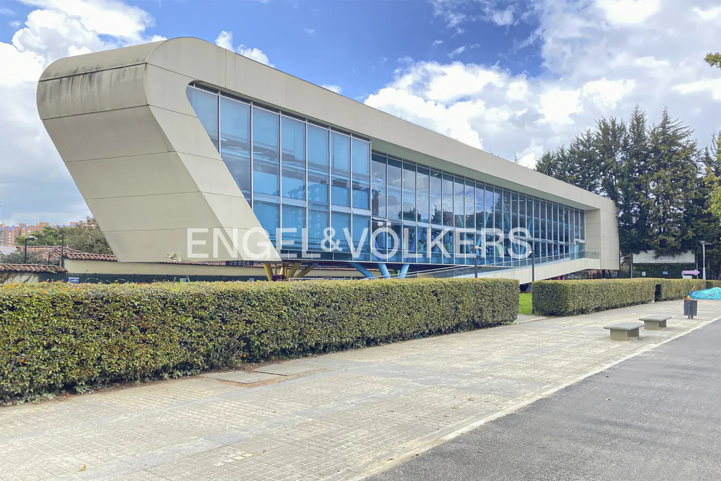 Modern, white building with blue-tinted windows and Engel & Völkers logo. Green hedges line the walkway. Blue sky with clouds.