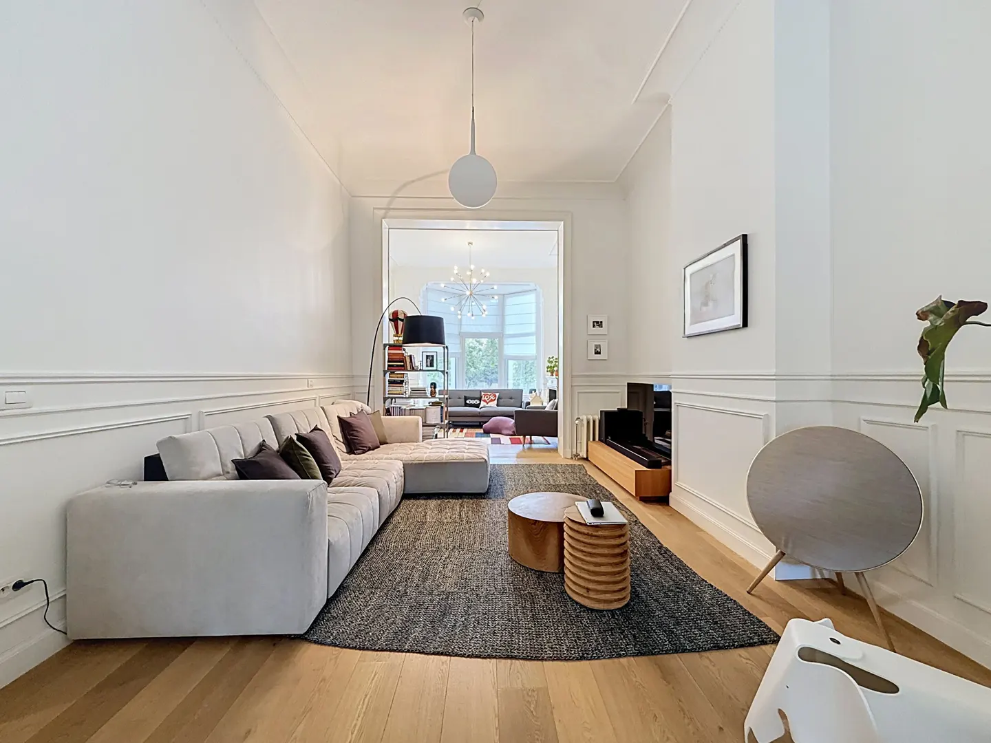 Bright living room with a large grey sectional sofa, dark area rug, wooden coffee tables, and a view into a second room.