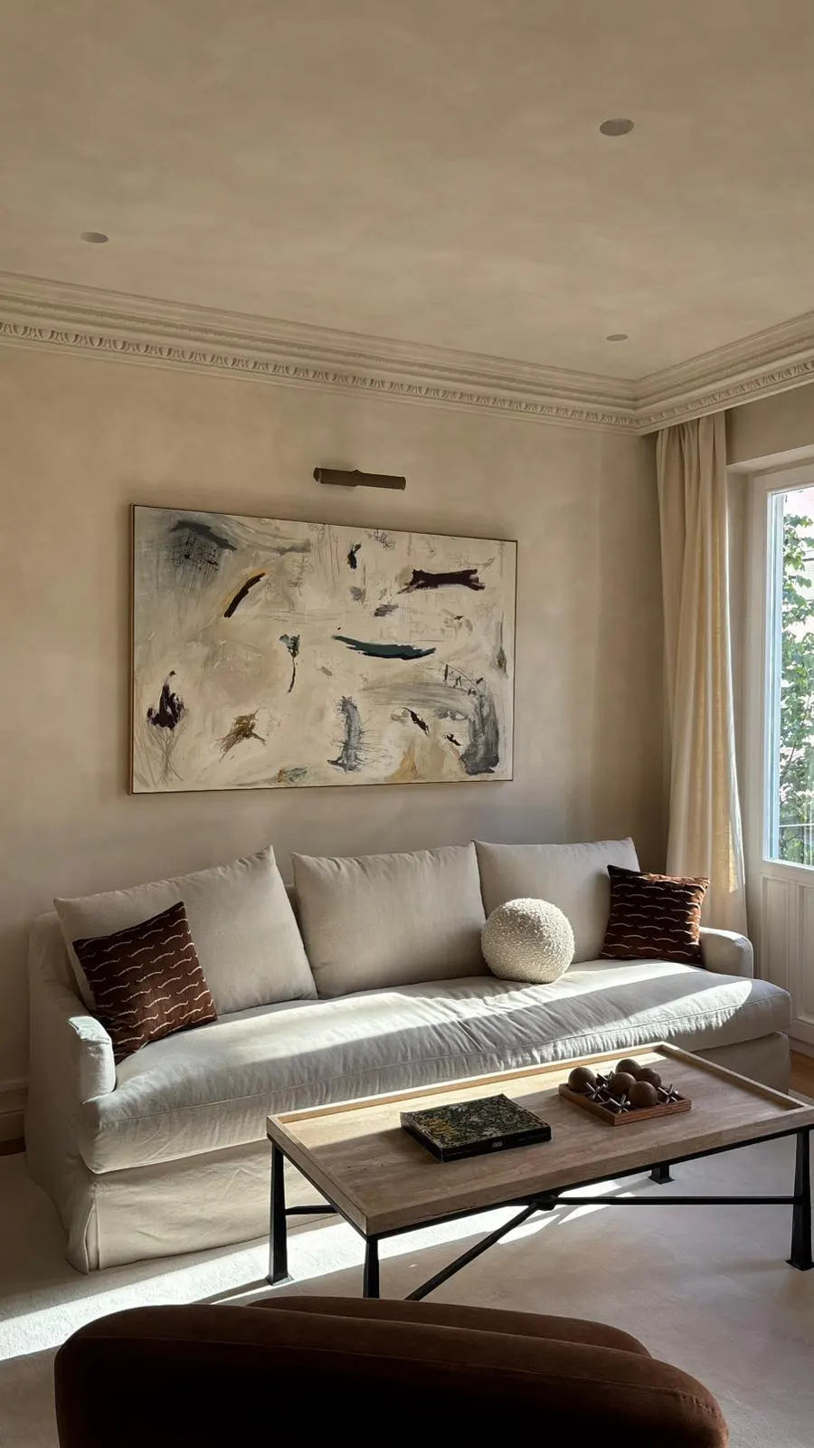 Living room with a beige sofa, brown pillows, and a wooden coffee table. An abstract painting hangs above the sofa. Cream walls and a window with white curtains.