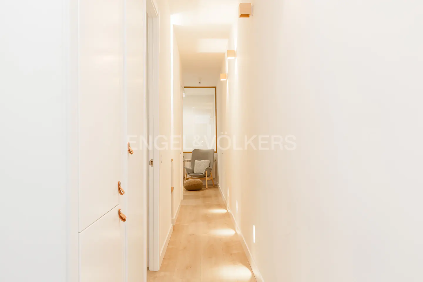 Bright hallway with white walls, wood floors, and recessed lighting. A gray chair and ottoman are visible at the end of the hall. Engel & Volkers logo.