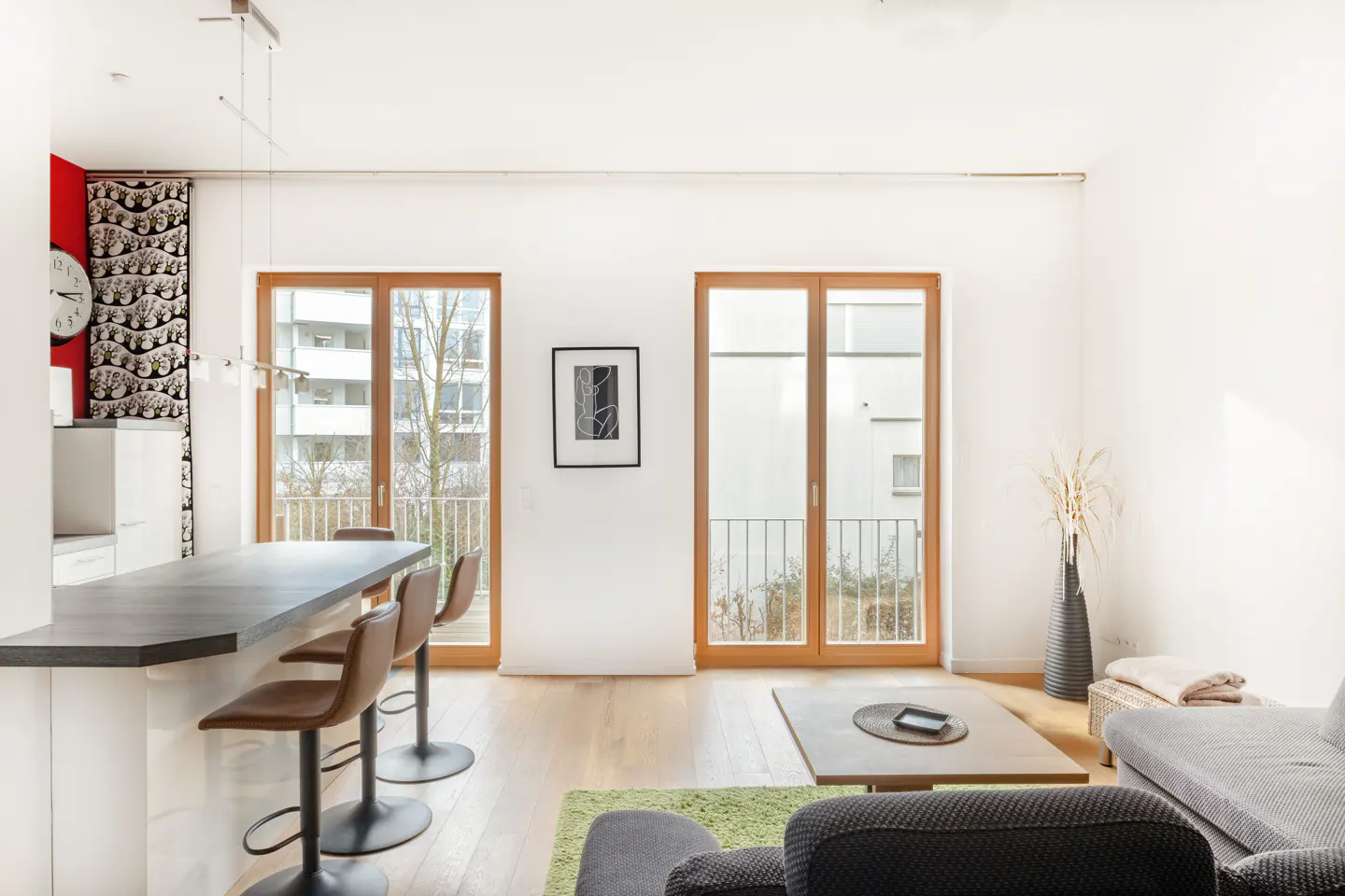 Bright, modern apartment interior with wood floors, bar stools at a counter, and sliding glass doors to balconies. A gray sofa is visible.