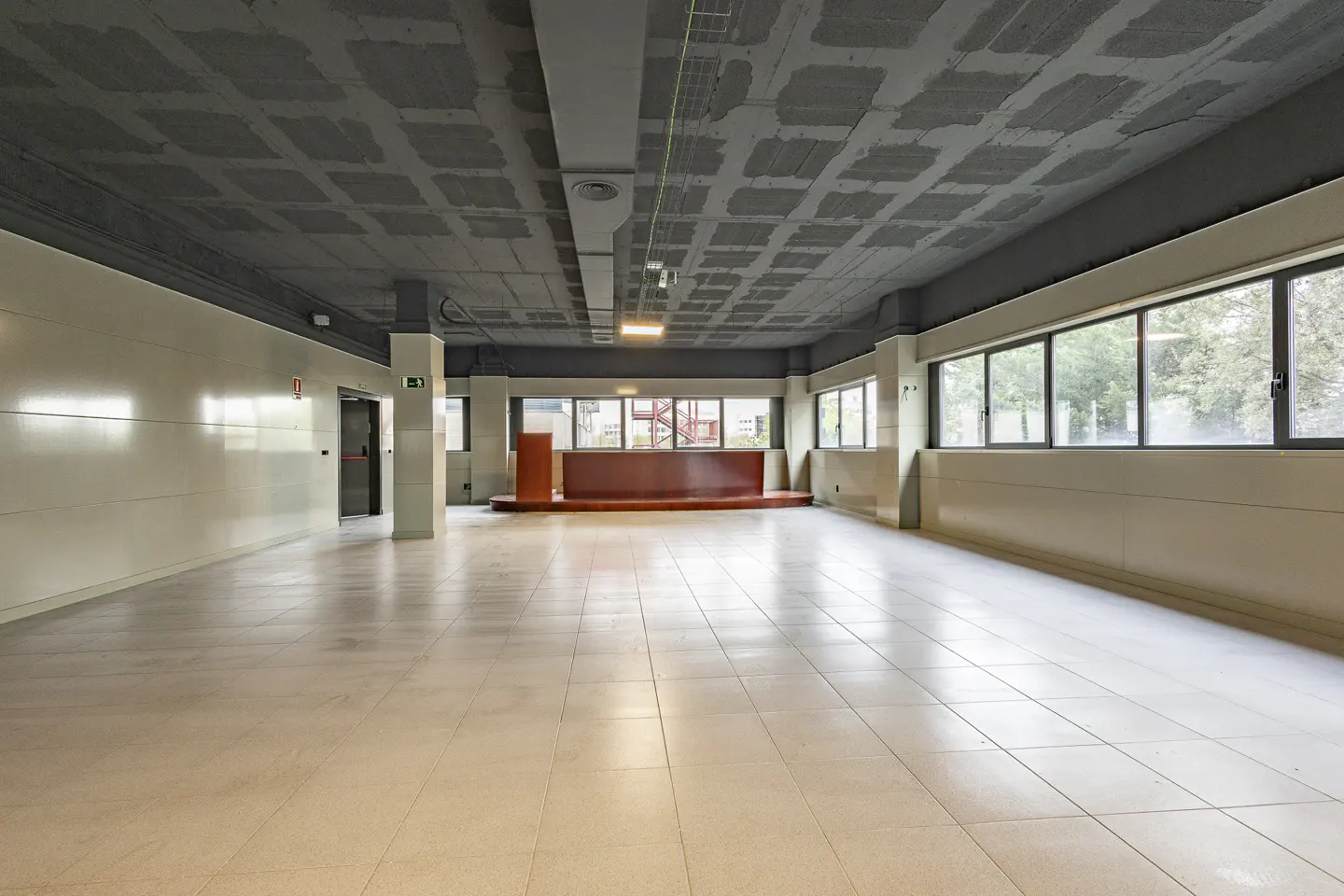 A large, empty room with white tile floors, a gray ceiling, and a brown stage area. Windows line one wall, offering a view of trees.