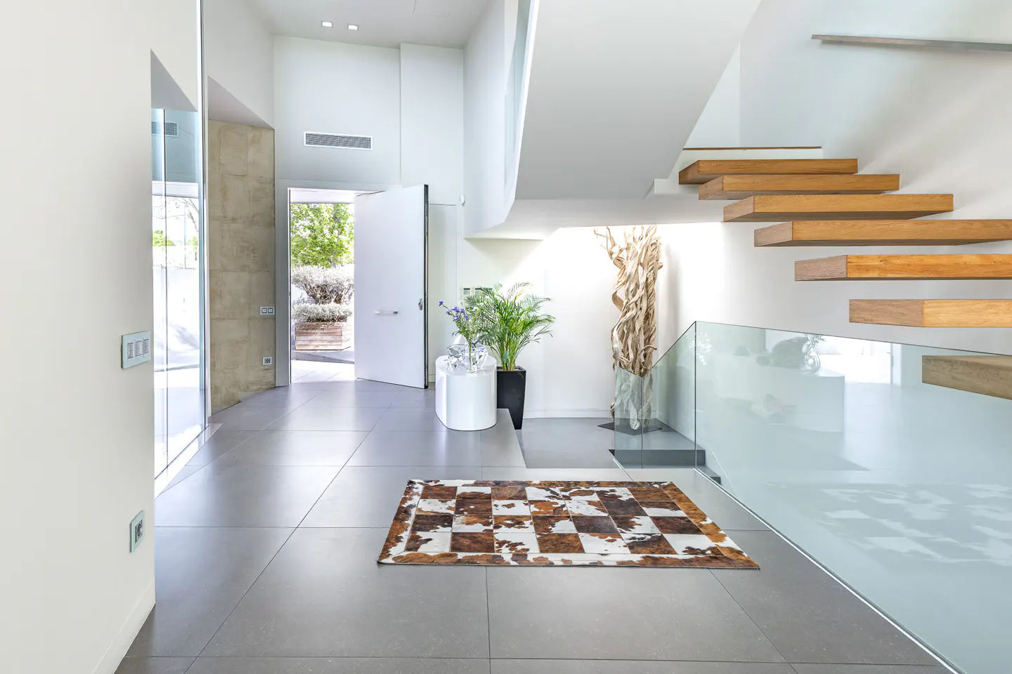 Bright foyer with gray tile floor, white walls, and floating wood stairs with glass railing. A brown and white rug sits near an open white door.