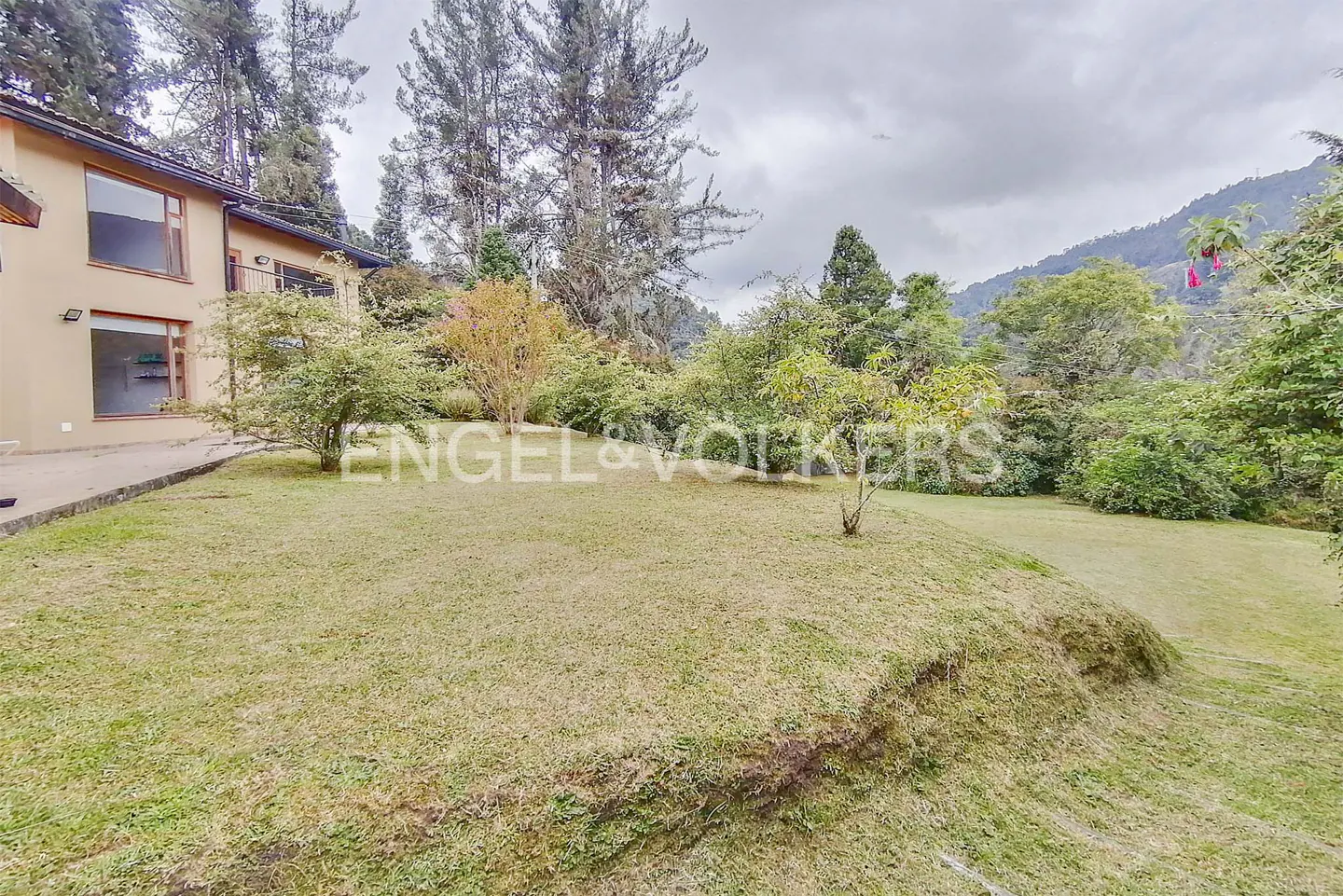 Exterior view of a tan house with a large green lawn, trees, and mountains in the background on a cloudy day.