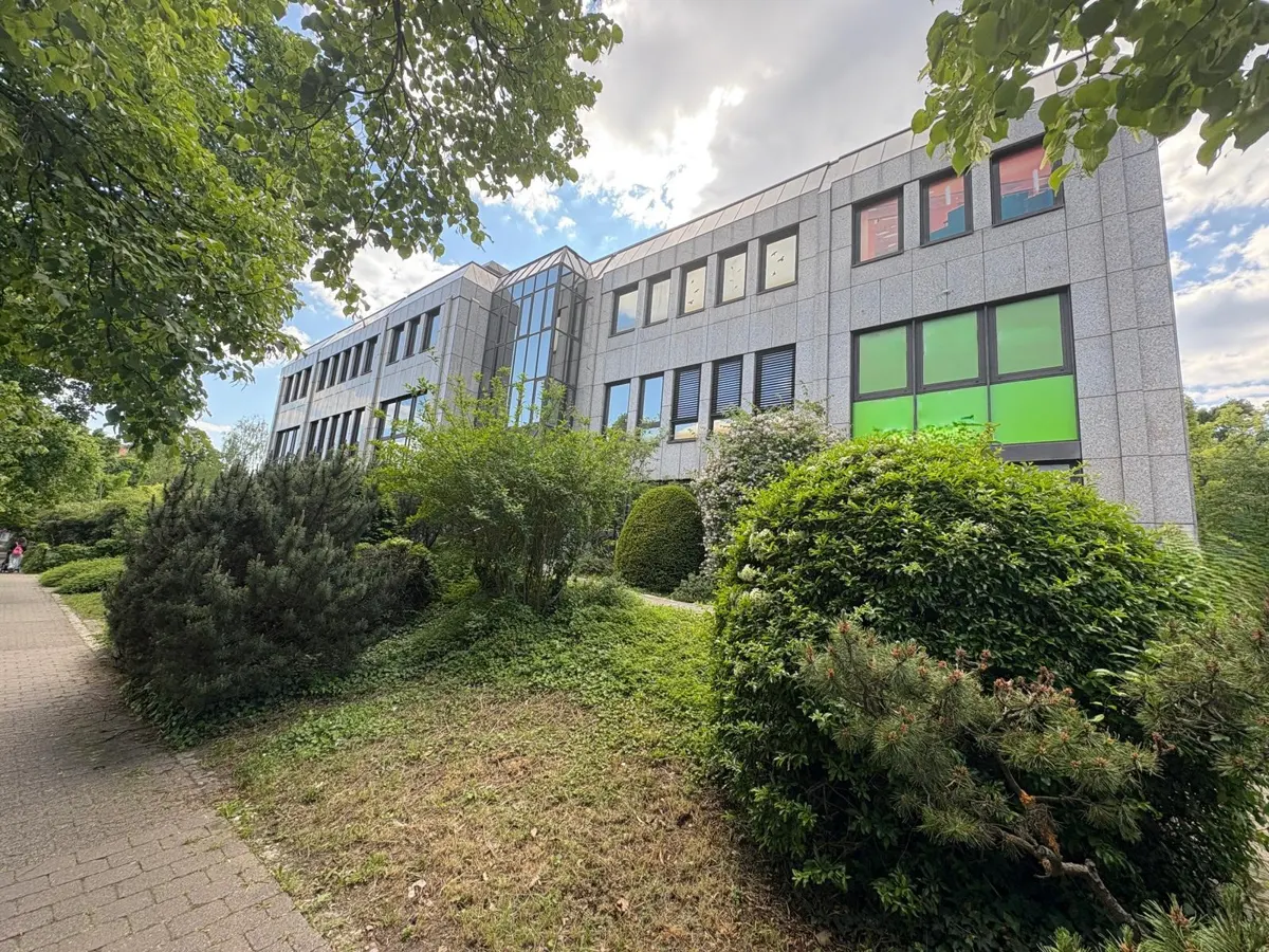 Exterior view of a two-story gray stone building with green and red window tints, surrounded by lush green landscaping.