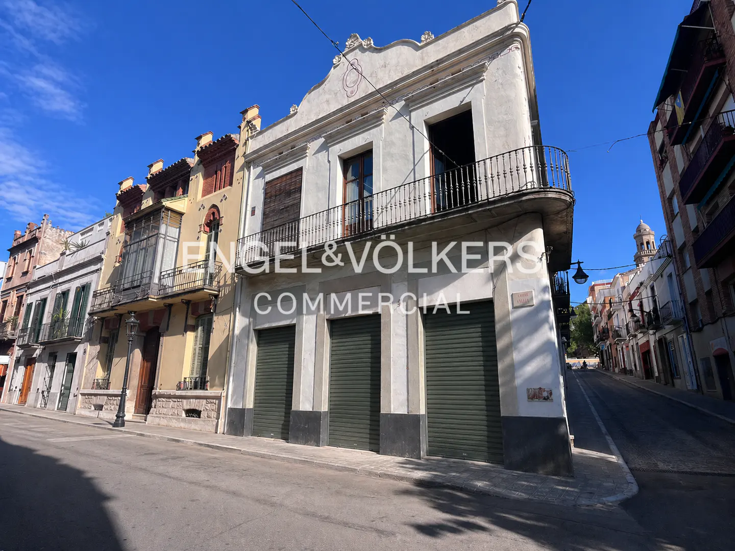 Street view of a two-story white building with green roll-up doors and a black iron balcony. Other buildings line the street under a blue sky.
