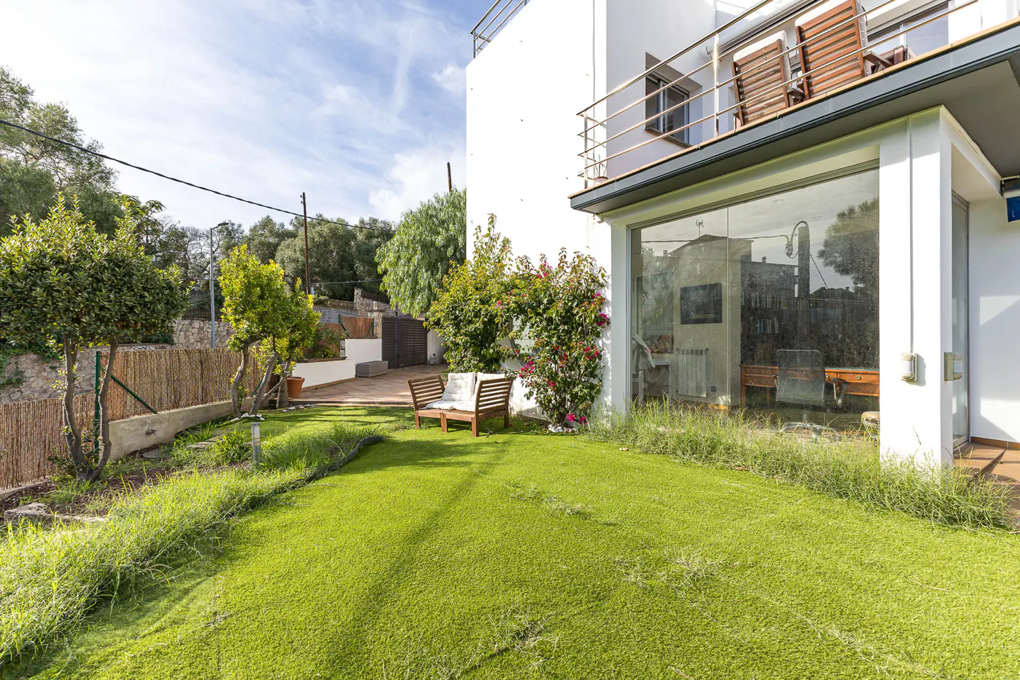 Exterior view of a modern white house with a green lawn, wooden bench, and a balcony with chairs under a blue sky.