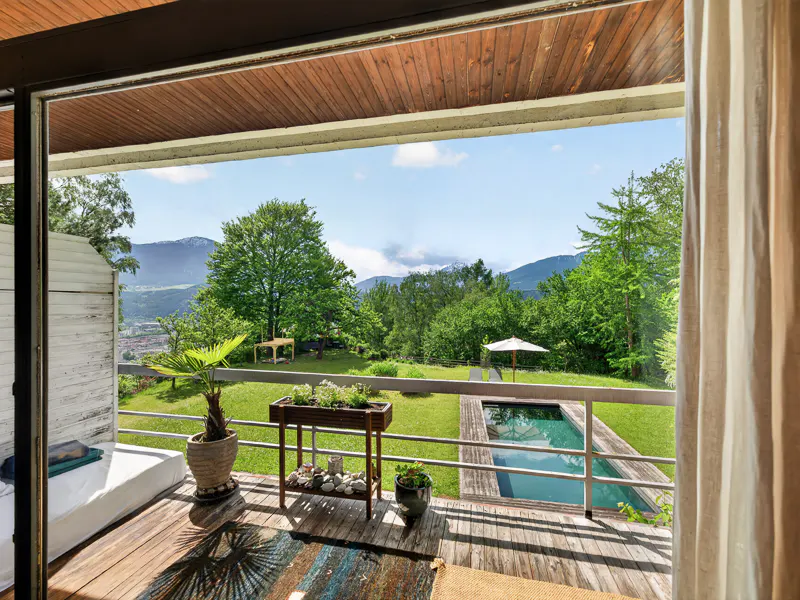 View from a wood-floored balcony with potted plants, overlooking a green lawn, pool, trees, and mountains under a blue sky.