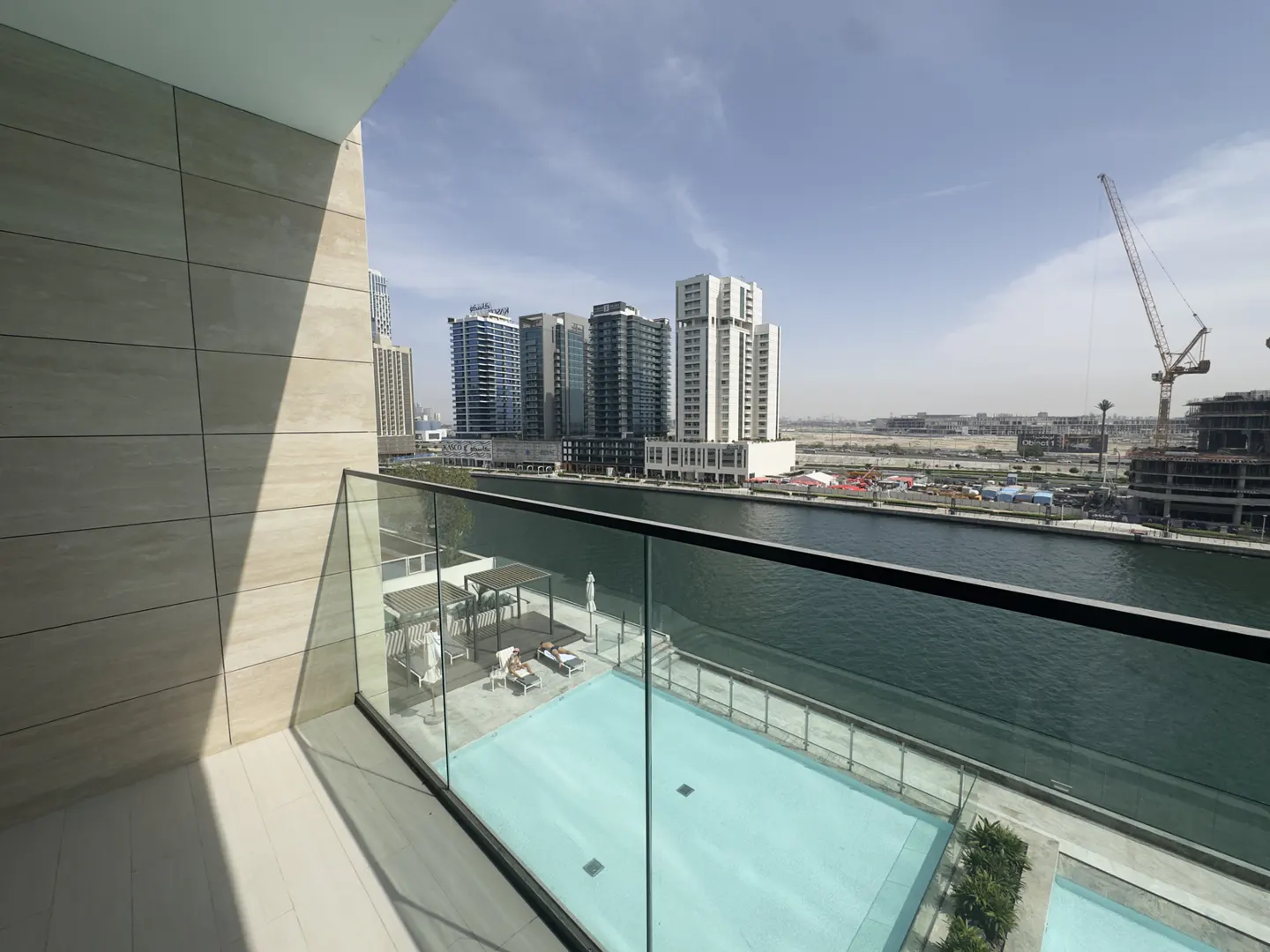 Balcony view of a pool, river, and city skyline. Modern buildings and a construction crane are visible in the background.