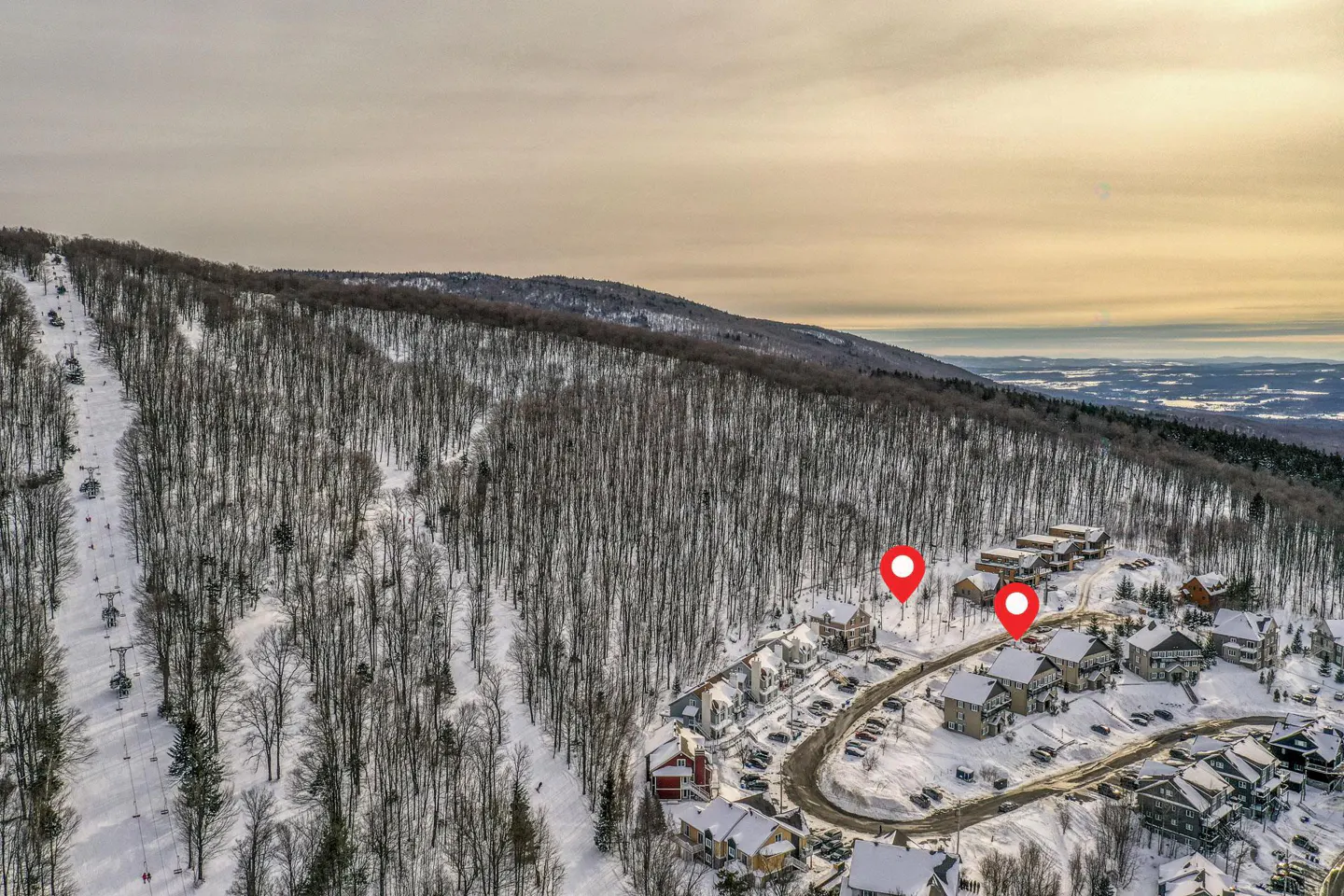 Aerial view of a ski resort with snow-covered slopes, trees, and chalets. Two red location pins mark properties.