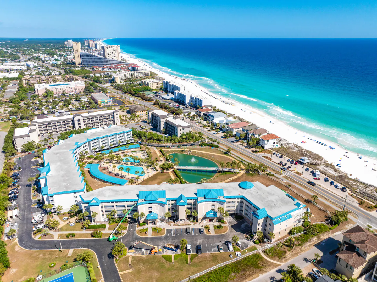 Aerial view of a beachfront resort with turquoise roofs, a pool, and a pond with a bridge, near a white sand beach and blue ocean.