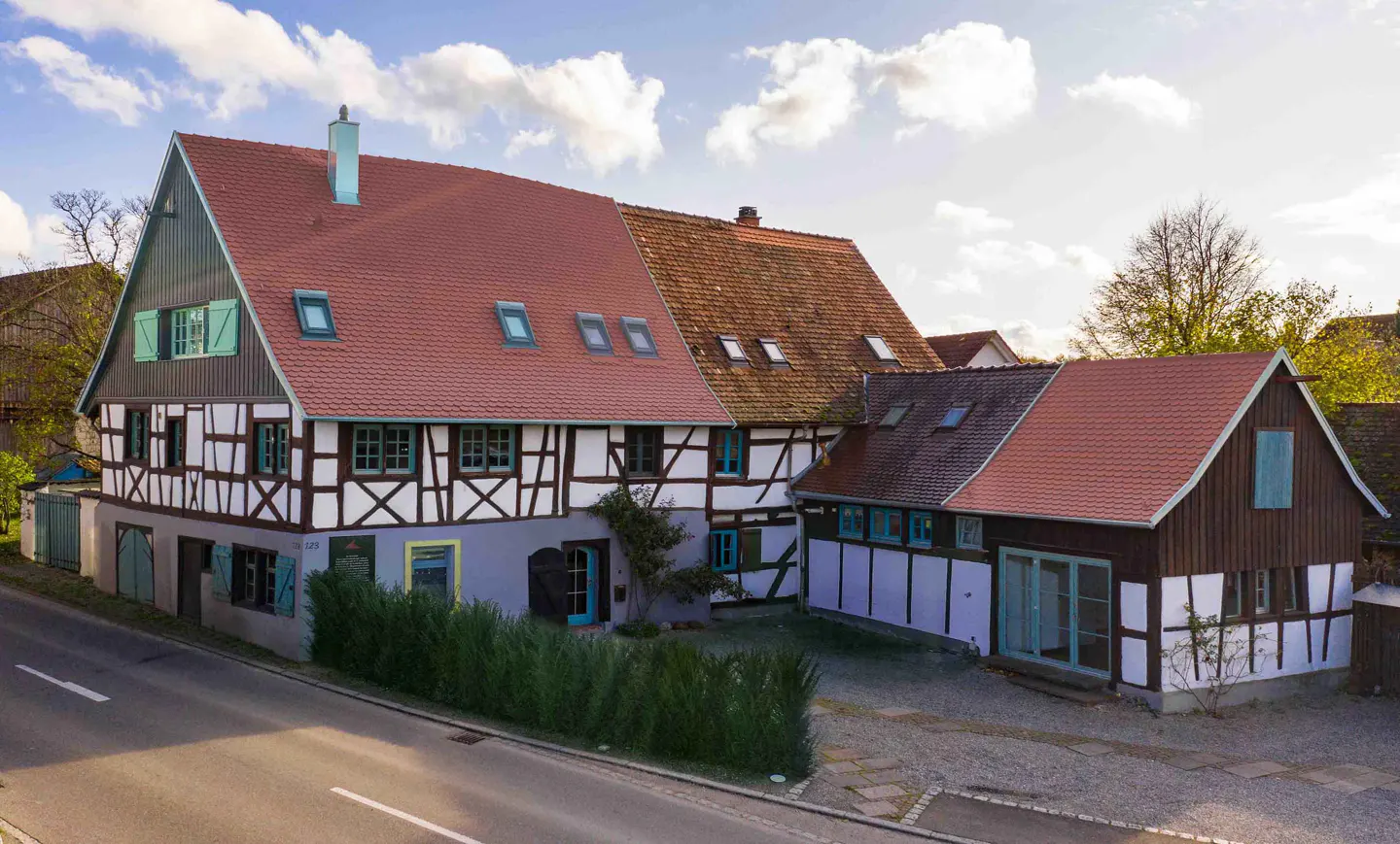 A traditional half-timbered house with a red tiled roof, blue shutters, and a gravel driveway next to a paved road.