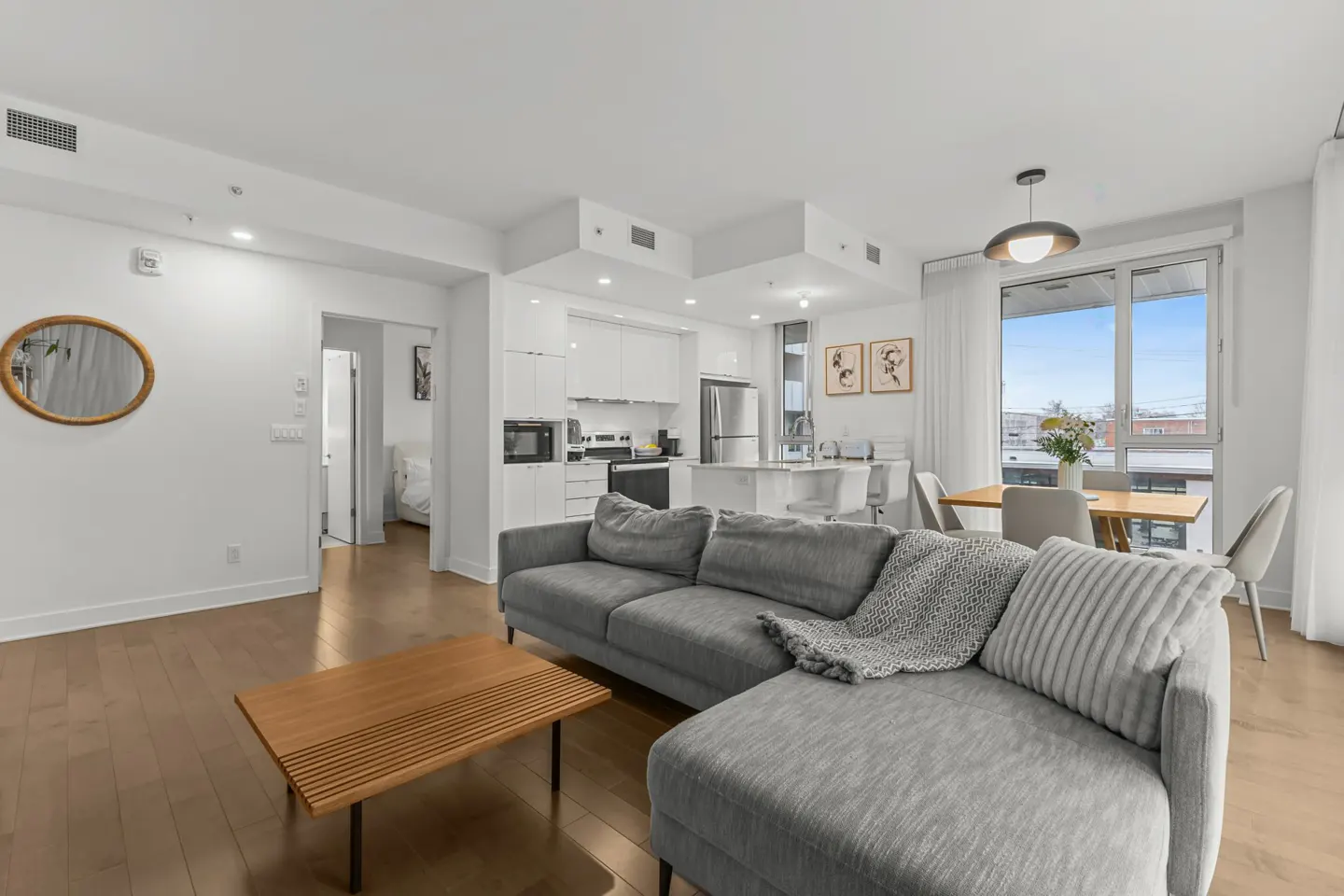 Bright, modern condo interior with a gray sectional sofa, wood floors, and a white kitchen. A dining table sits near a large window.