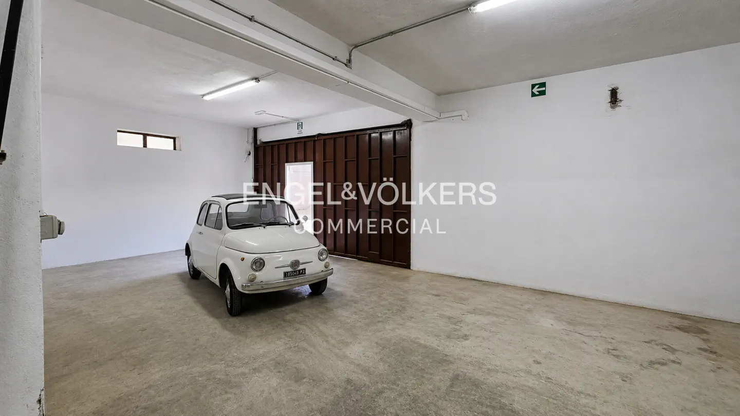 A white Fiat 500 parked in a large, empty garage with white walls and a concrete floor. A brown metal door is in the background.