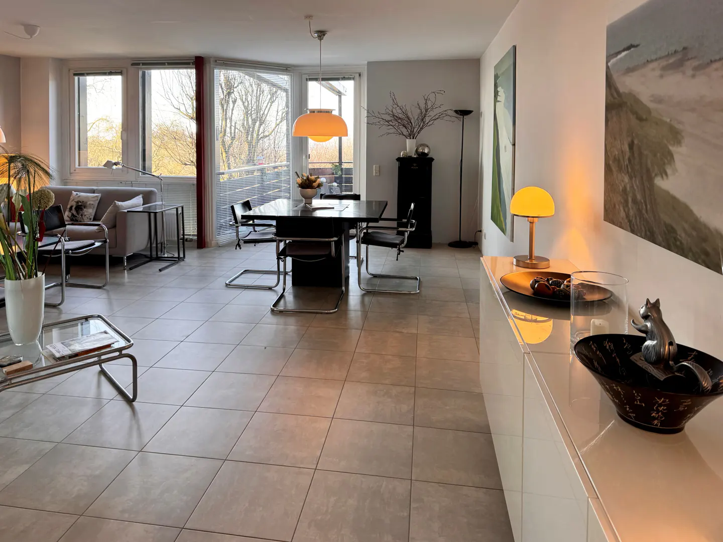 A modern apartment interior with a dining table, sofa, and white sideboard. Art adorns the walls, and natural light streams through large windows.