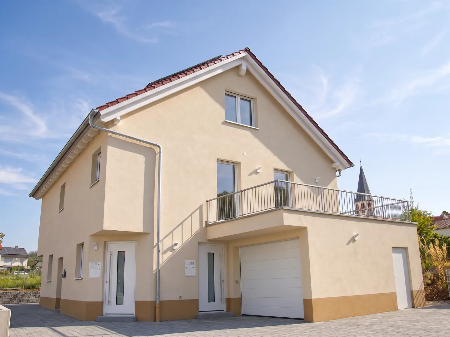 Two-story beige house with a red roof, white trim, and a balcony above the garage door. Blue sky in the background.