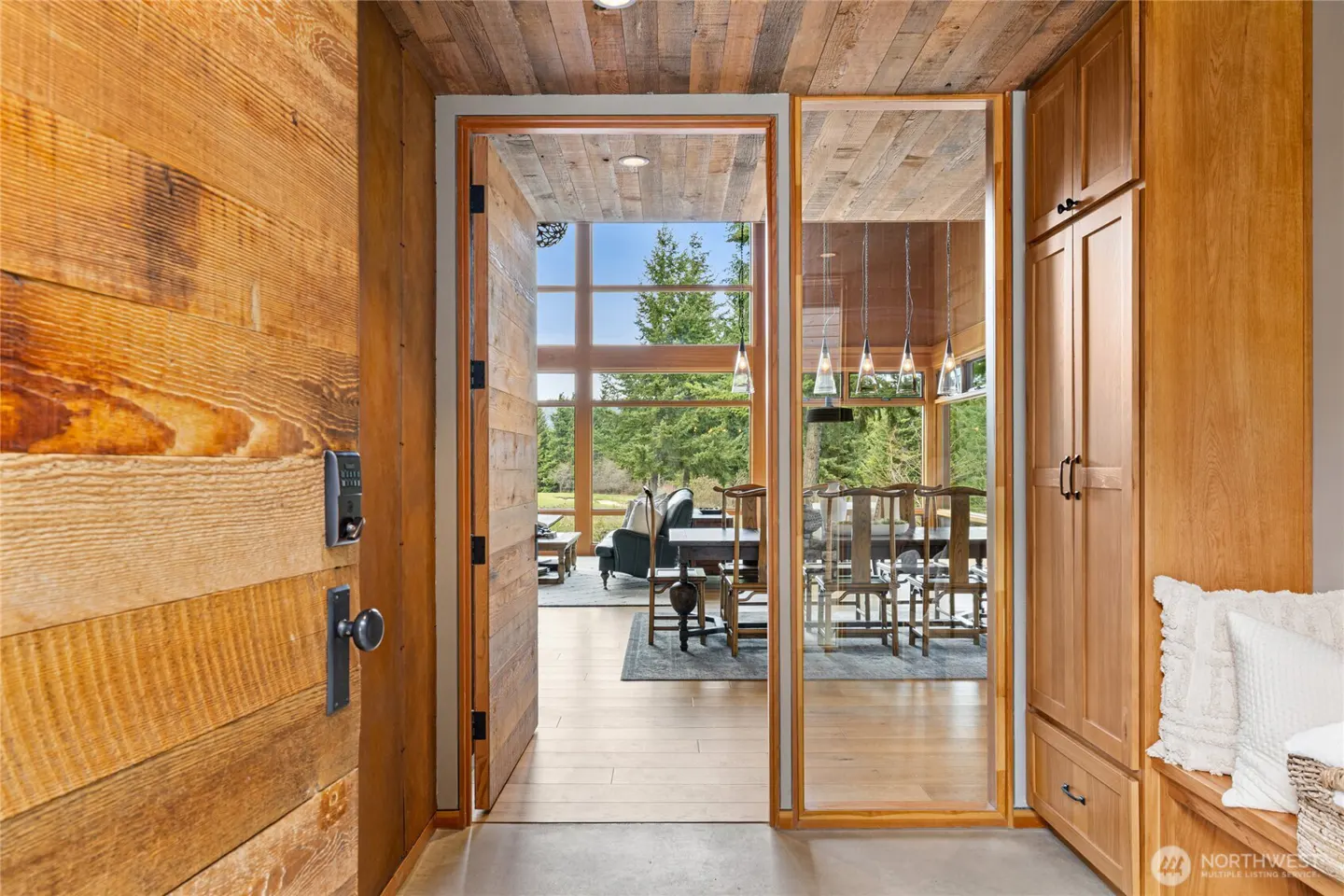 Open wood door reveals a modern home interior with a dining table, living room, and large windows overlooking a green landscape.