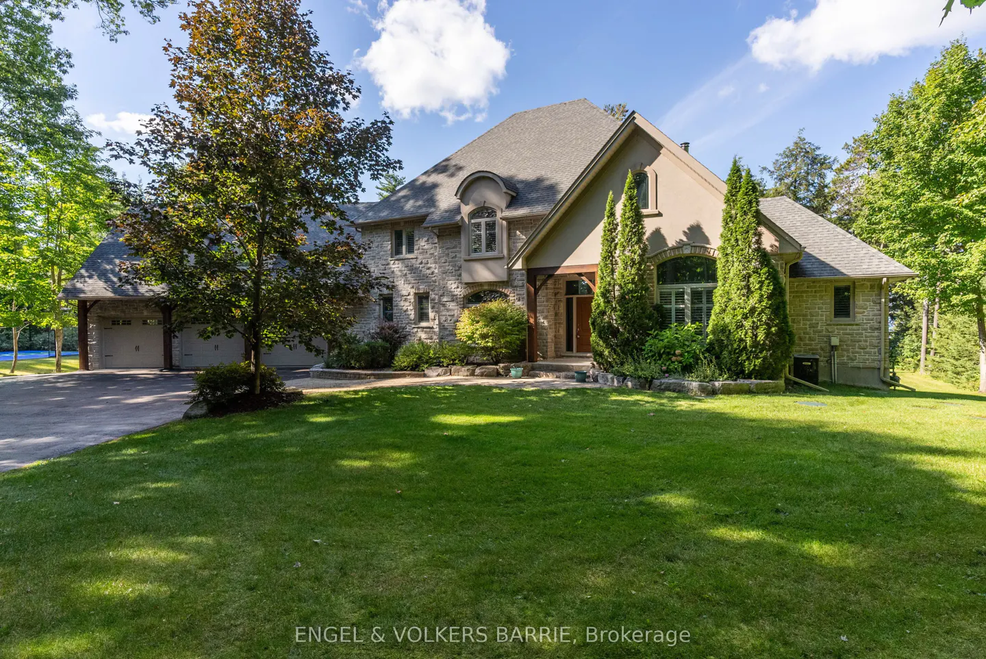 Exterior view of a large, two-story stone and stucco house with a gray roof and a green lawn. Trees surround the property.